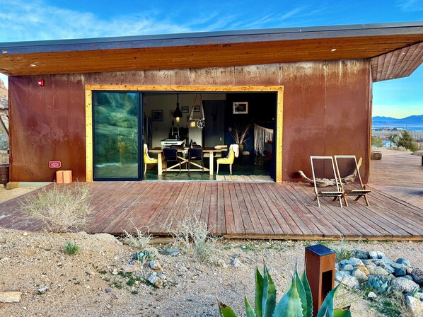 Un bâtiment moderne doté d’une terrasse en bois, d’un coin repas avec des chaises et une table, et offrant une vue sur le paysage.