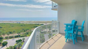 A balcony with a view of the ocean and a road. There are two blue chairs and a small table.