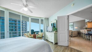 A hotel room with a large bed, a ceiling fan, a dresser, and a view of the cityscape through large windows.