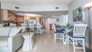 A spacious living area with a sofa, dining table, and chairs, a kitchen in the background, and a colorful abstract painting on the wall.