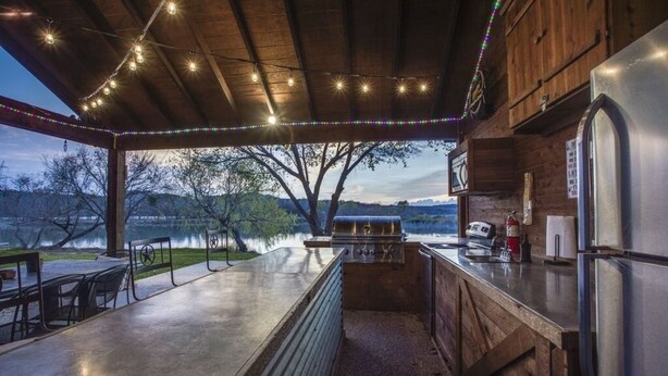 A covered outdoor kitchen with a grill, refrigerator, and dining area overlooking a lake.