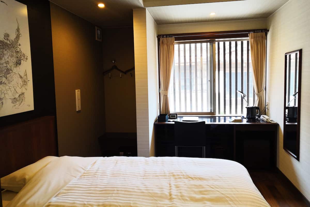 A hotel room with a bed, a desk with a chair, a window with curtains, and an air conditioning unit.