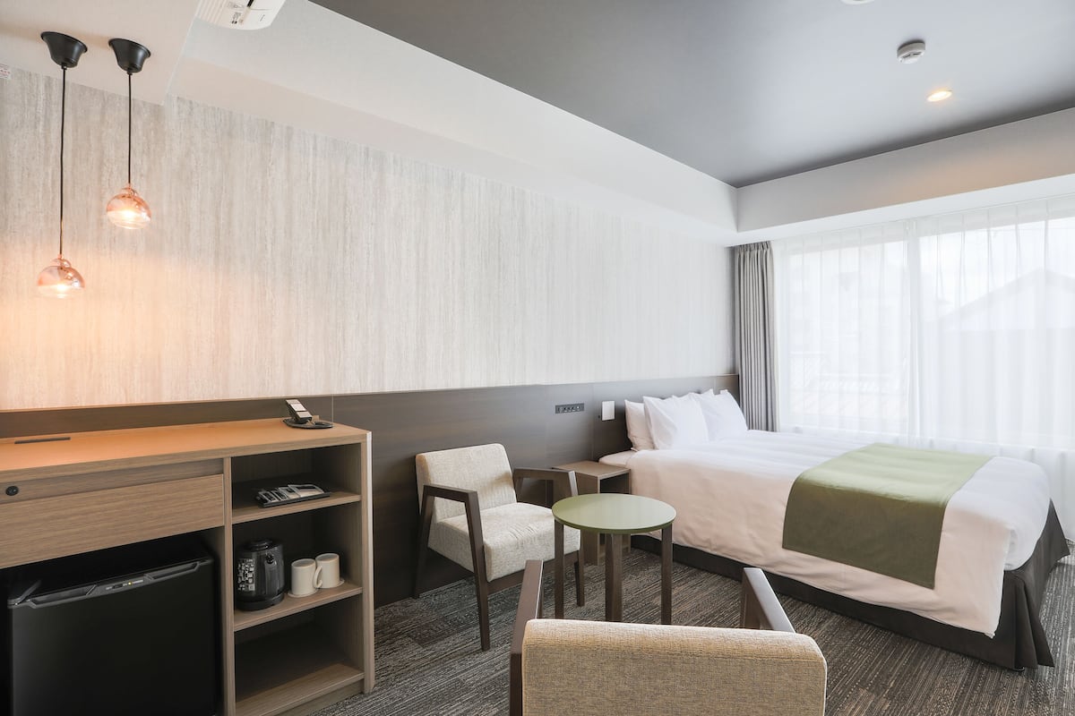 A hotel room with a bed, a desk, a chair, a small table, and a coffee machine.
