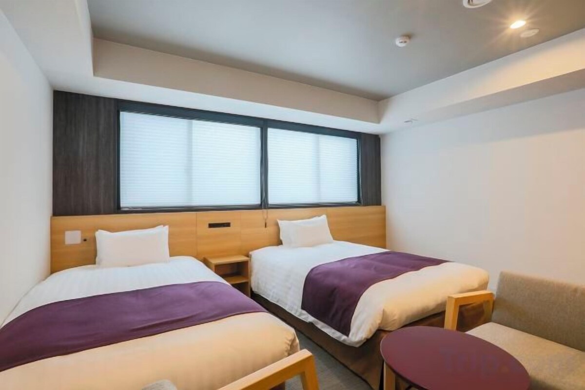 A hotel room with two beds, a sofa, a small table, and a window with blinds.