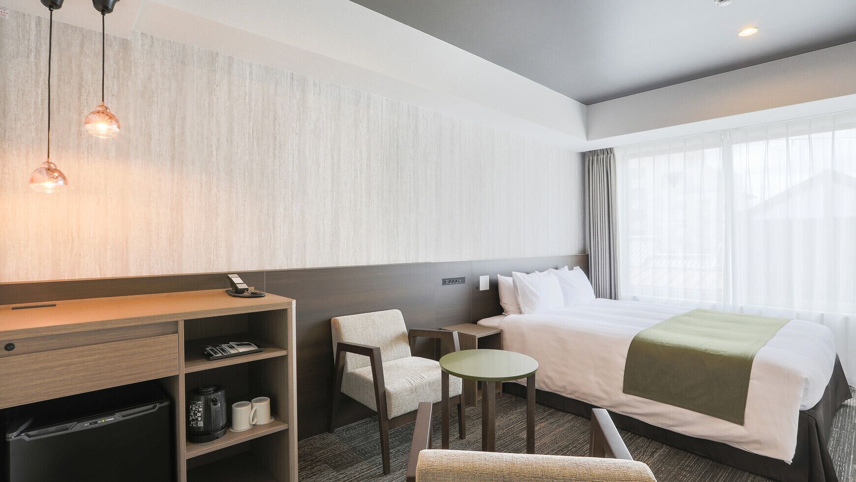 A hotel room with a bed, a desk, a chair, a small table, and a wall-mounted shelf.