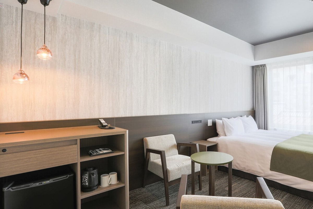 A hotel room with a bed, a desk, a chair, a small table, and a wall-mounted shelf.