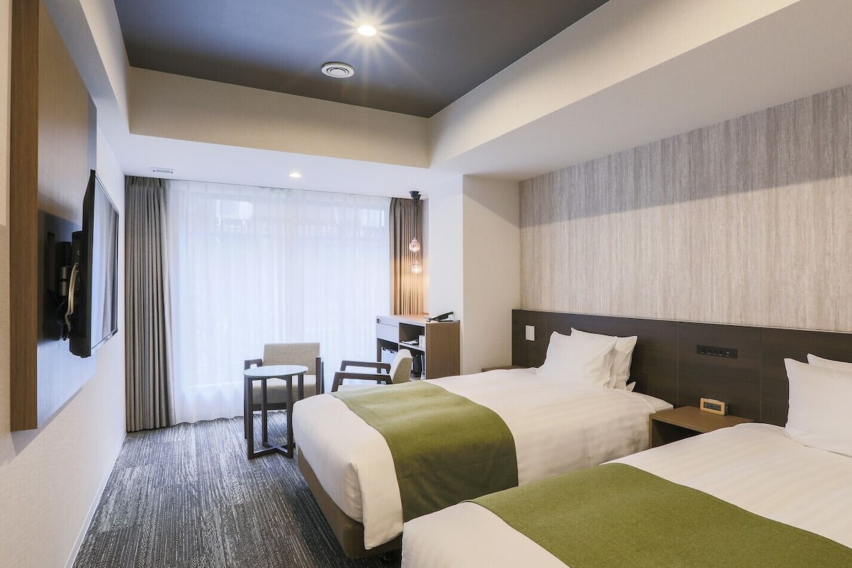 A hotel room with two beds, a desk, a chair, a TV, and a large window with curtains.