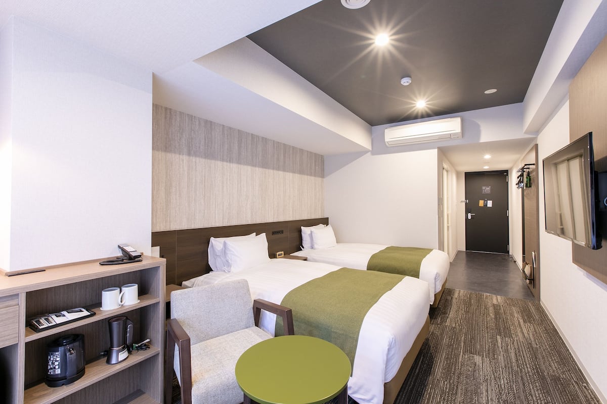 A hotel room with two beds, a small table, a chair, a shelf with a coffee maker, and a TV.