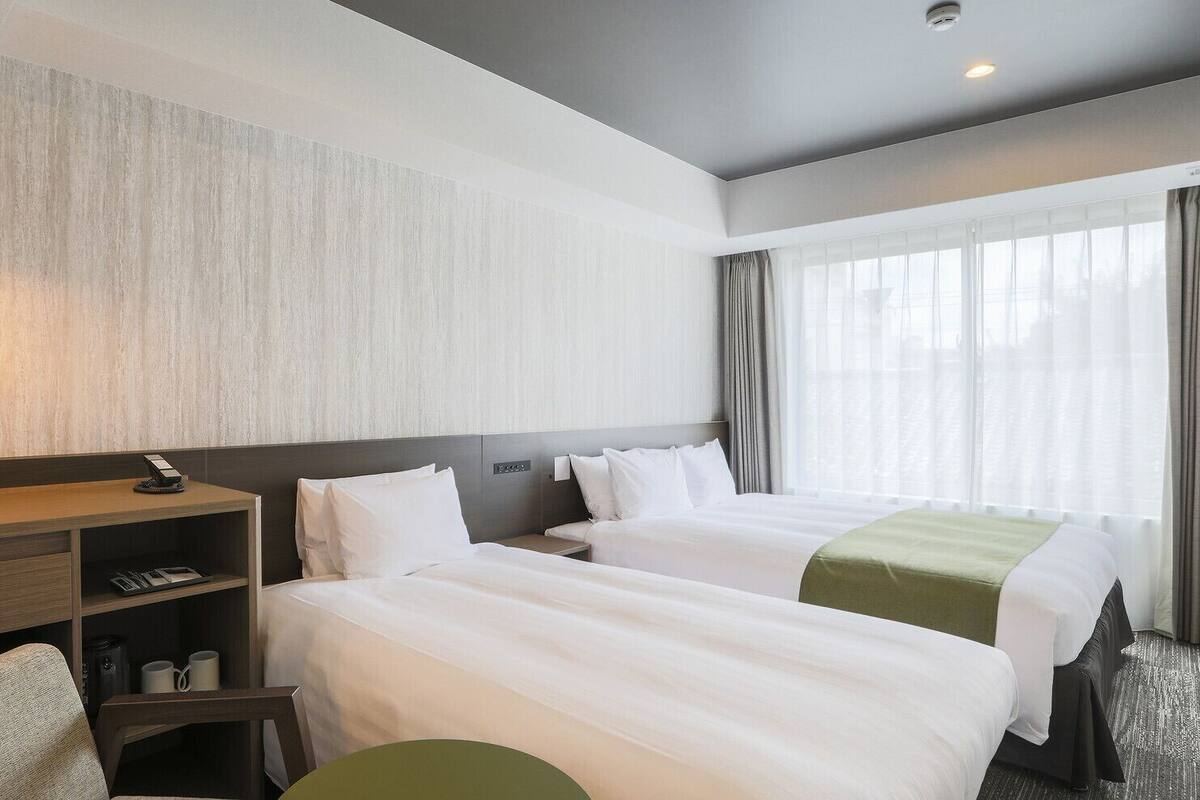 A hotel room with two beds, a desk, a chair, and a large window with curtains.