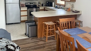 A modern kitchen with wooden cabinets, a stainless steel refrigerator, a microwave, and a coffee maker. There is a dining area with wooden chairs and a table. - Beautiful Broadkill Beach home with a short walk to the beach. (Milton)