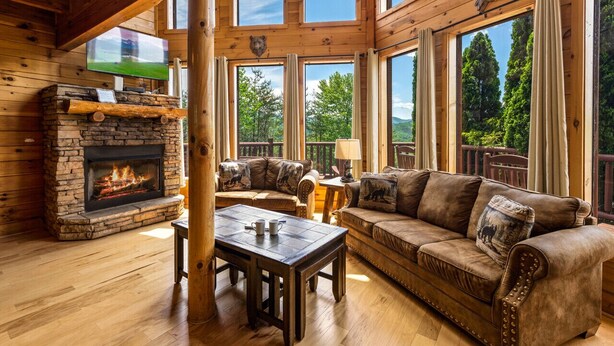 A cozy cabin interior with a stone fireplace, leather sofa, wooden floors, and large windows offering a view of the forest. - Luxury cabin w/indoor pool, hot tub, view, game rm (Sevierville)