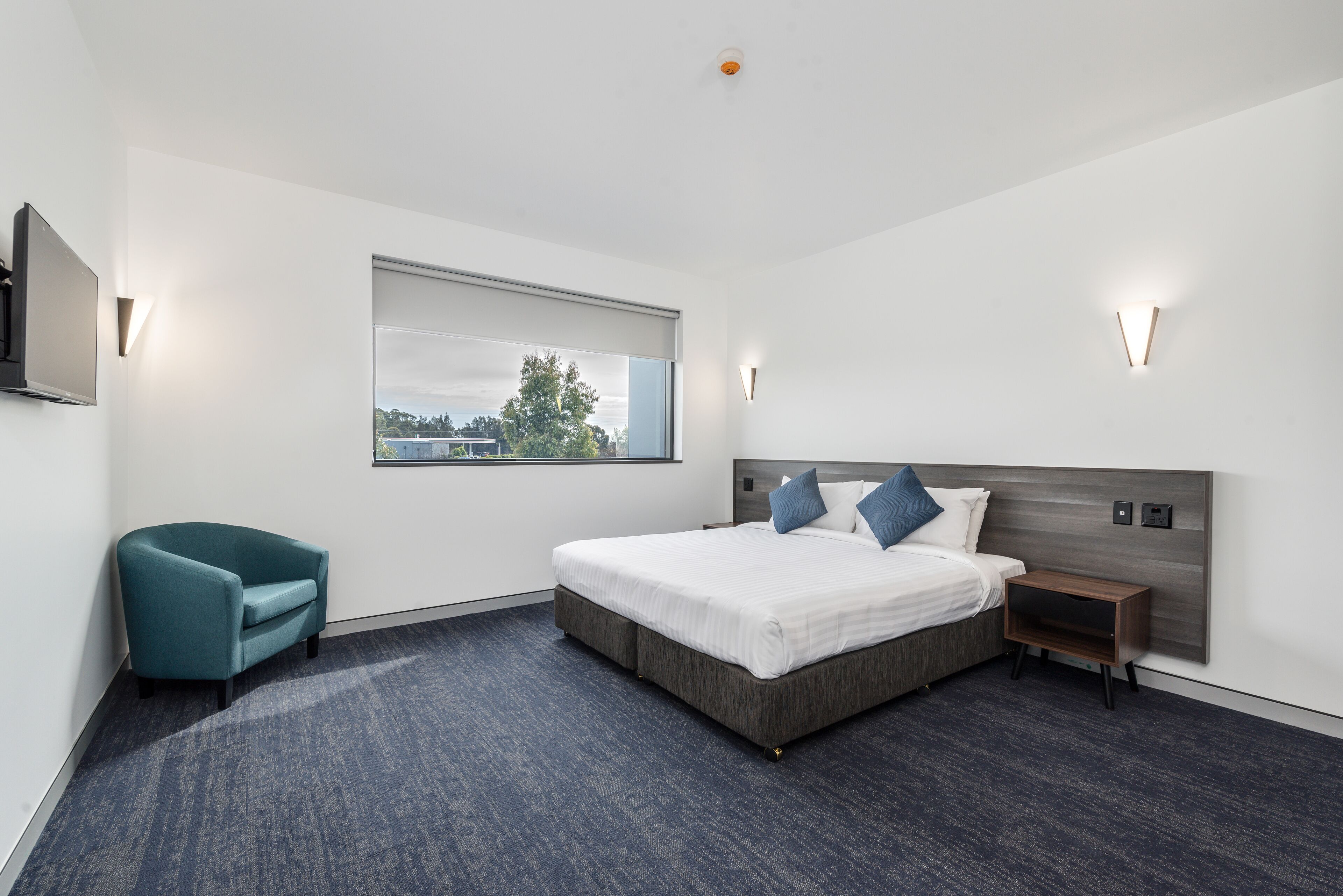Una habitación de hotel moderna con una cama grande, un sillón curvo color turquesa, una mesita de noche y una ventana con vistas a la ciudad.