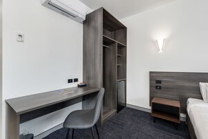 Habitación de hotel con escritorio, silla, armario, cama, mesita de noche y lámpara de pared.