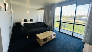 Una camera d'albergo con un letto, un divano nero, un tavolino in legno e ampie finestre con vista sull'esterno.