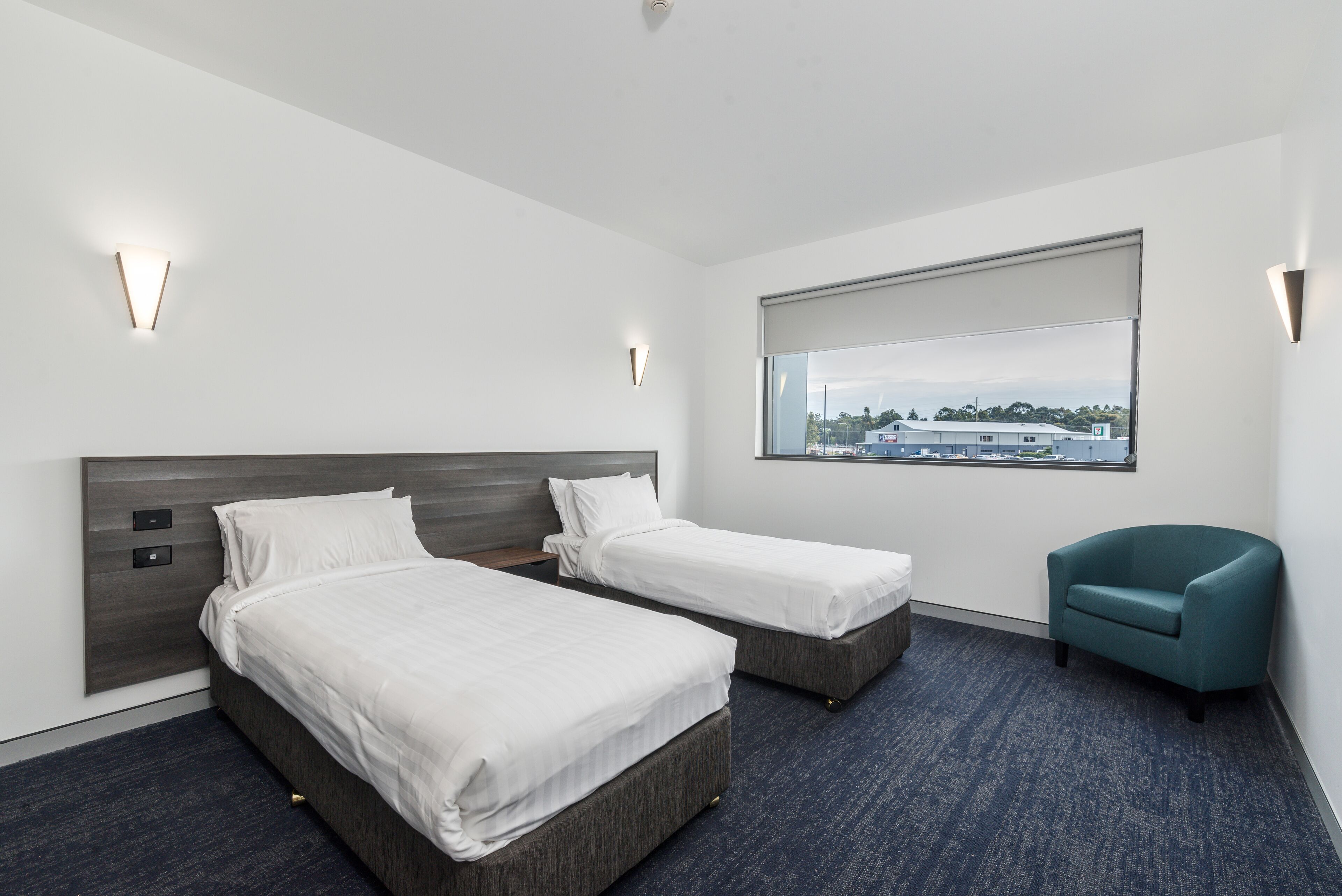 Quarto de hotel com duas camas, janela com vista e uma cadeira azul-esverdeada.