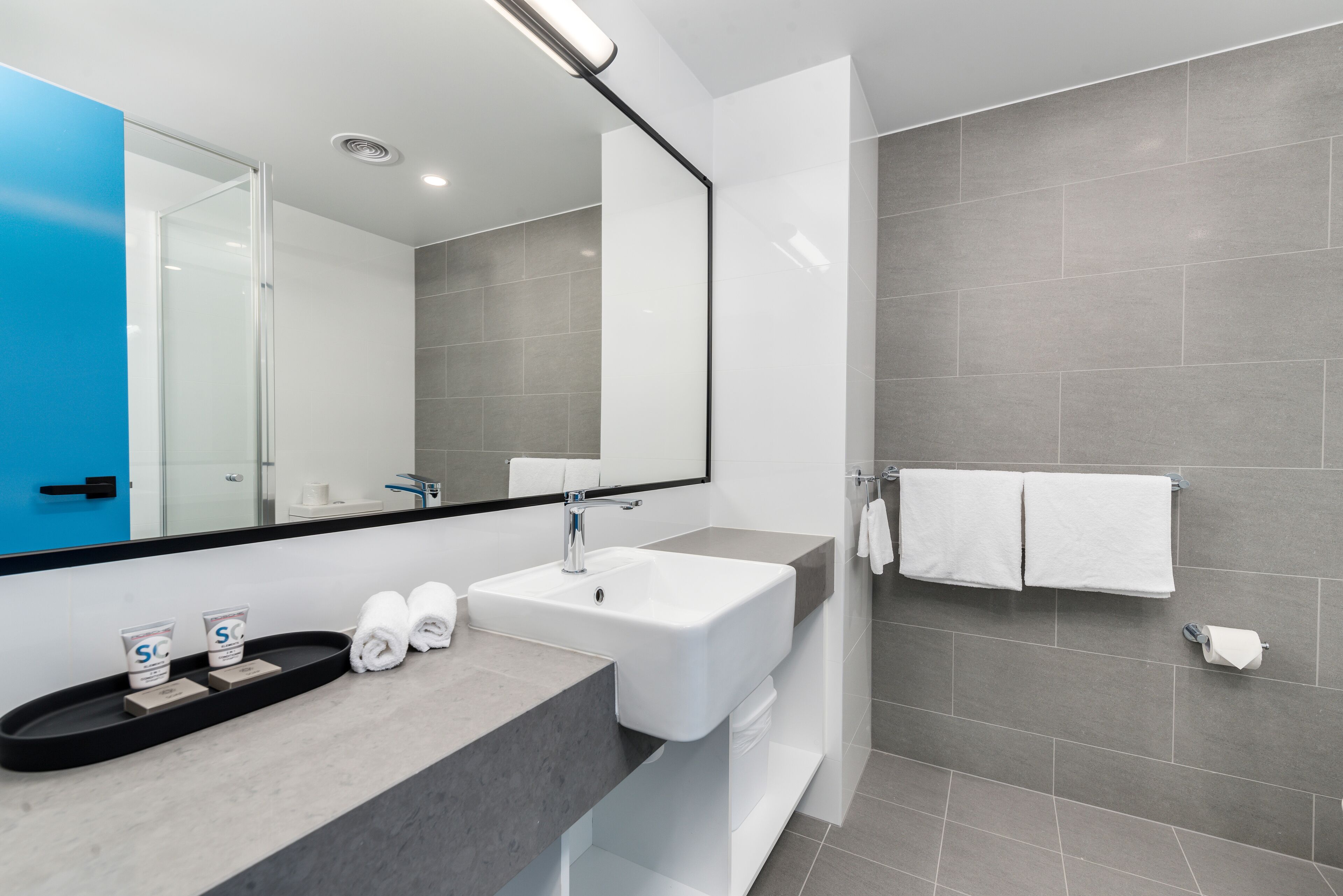 A modern bathroom with a large mirror, white sink, and grey tiles. There are two towels on the counter and a shower area with a glass door.
