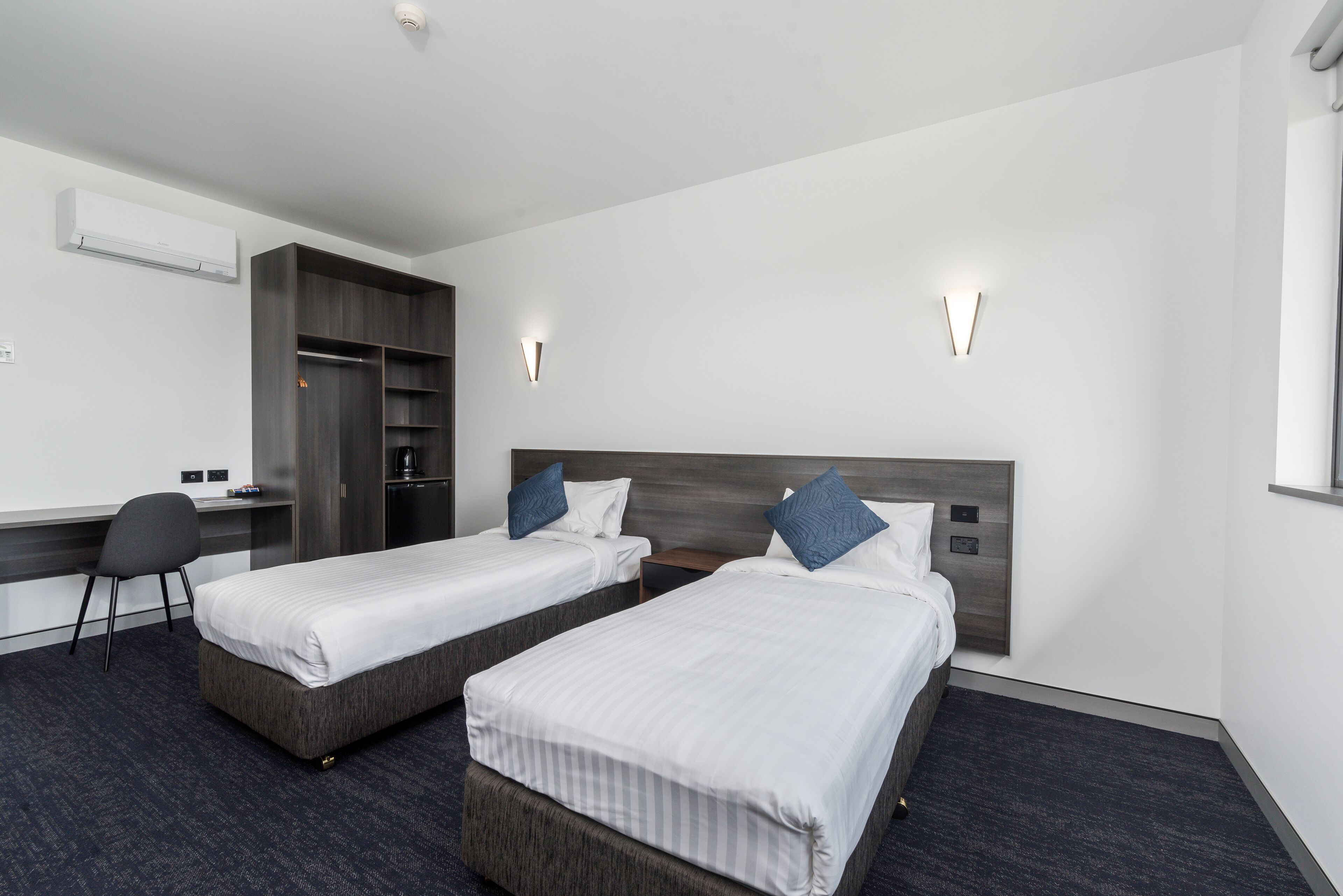 Een hotelkamer met twee enkele bedden, een bureau, een stoel, een kledingkast en een wandairconditioner.