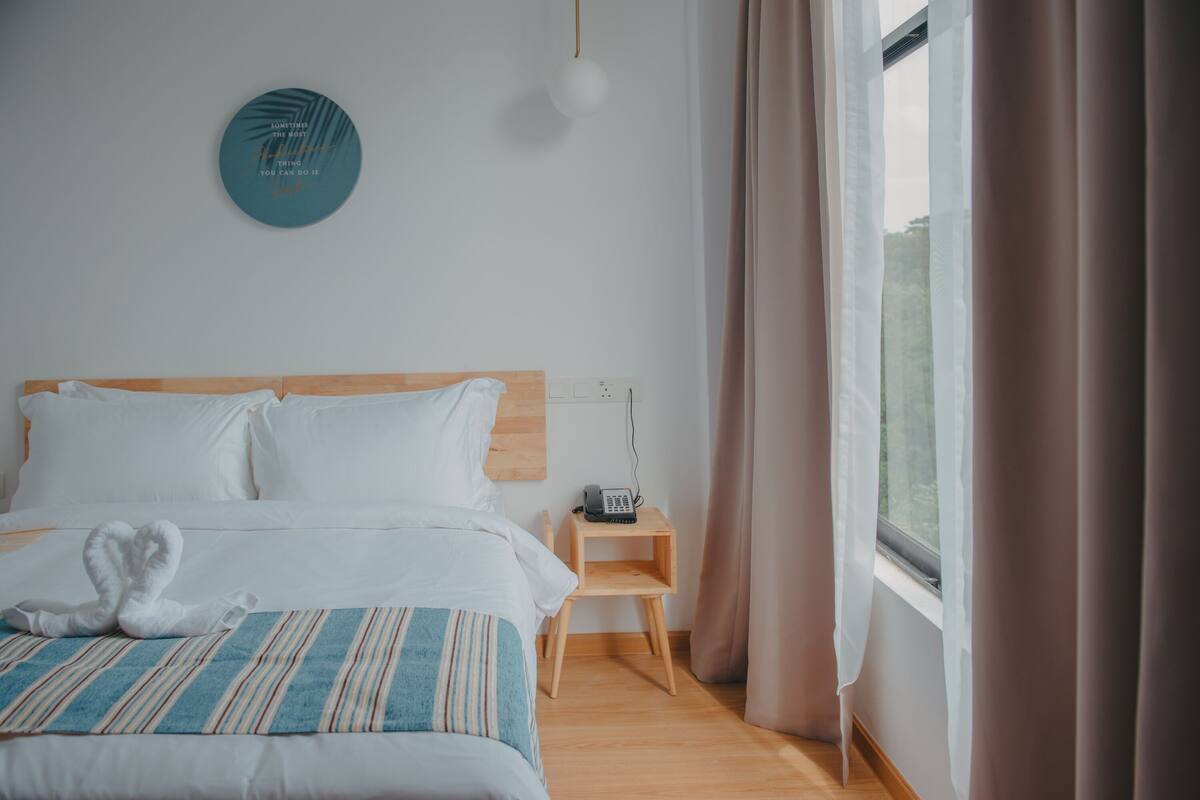 Een hotelkamer met een bed, nachtkastje met een lamp, een telefoon aan de muur en uitzicht op het landschap door een raam met gordijnen.