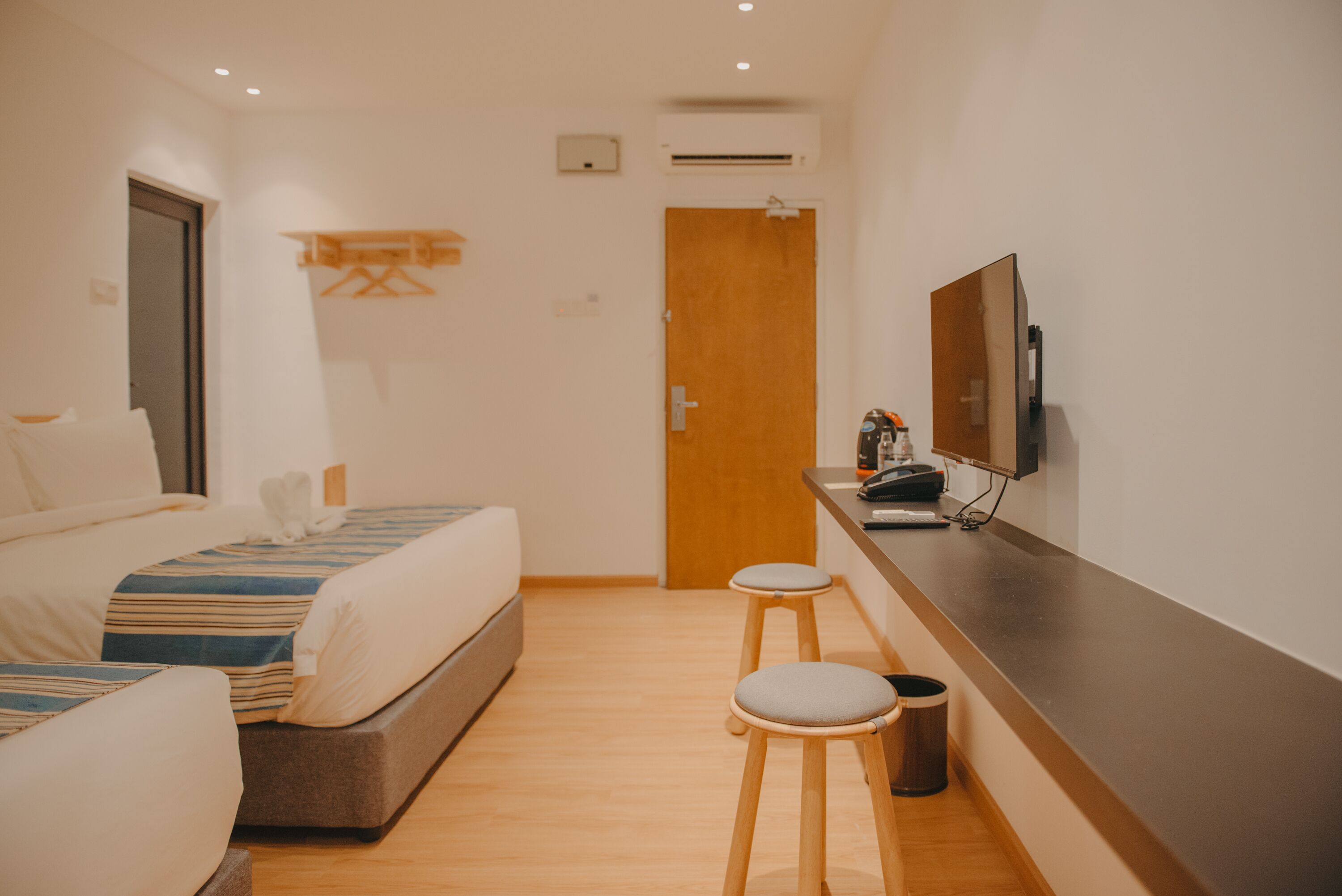 A hotel room with two beds, a wooden floor, a desk with a computer, and a TV mounted on the wall.
