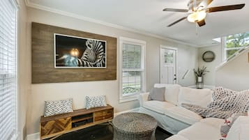 Un salon moderne avec un ventilateur de plafond, une grande œuvre d’art encadrée, un canapé blanc, un banc en bois et une petite table ronde.