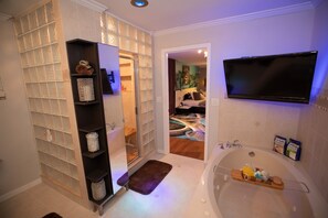 A modern bathroom with a glass shower enclosure, a bathtub with toiletries, and a flat-screen TV mounted above the tub.