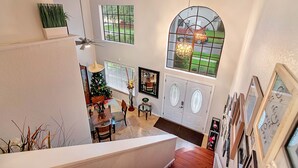 Amplia sala de estar con techo alto, una mesa de comedor, sillas y un árbol de Navidad. Hay varias ventanas y un gran ventanal de arco con una araña.