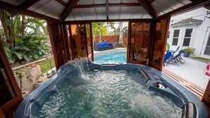 Un jacuzzi con techo de madera y vista a un patio trasero con piscina, sillas y plantas.