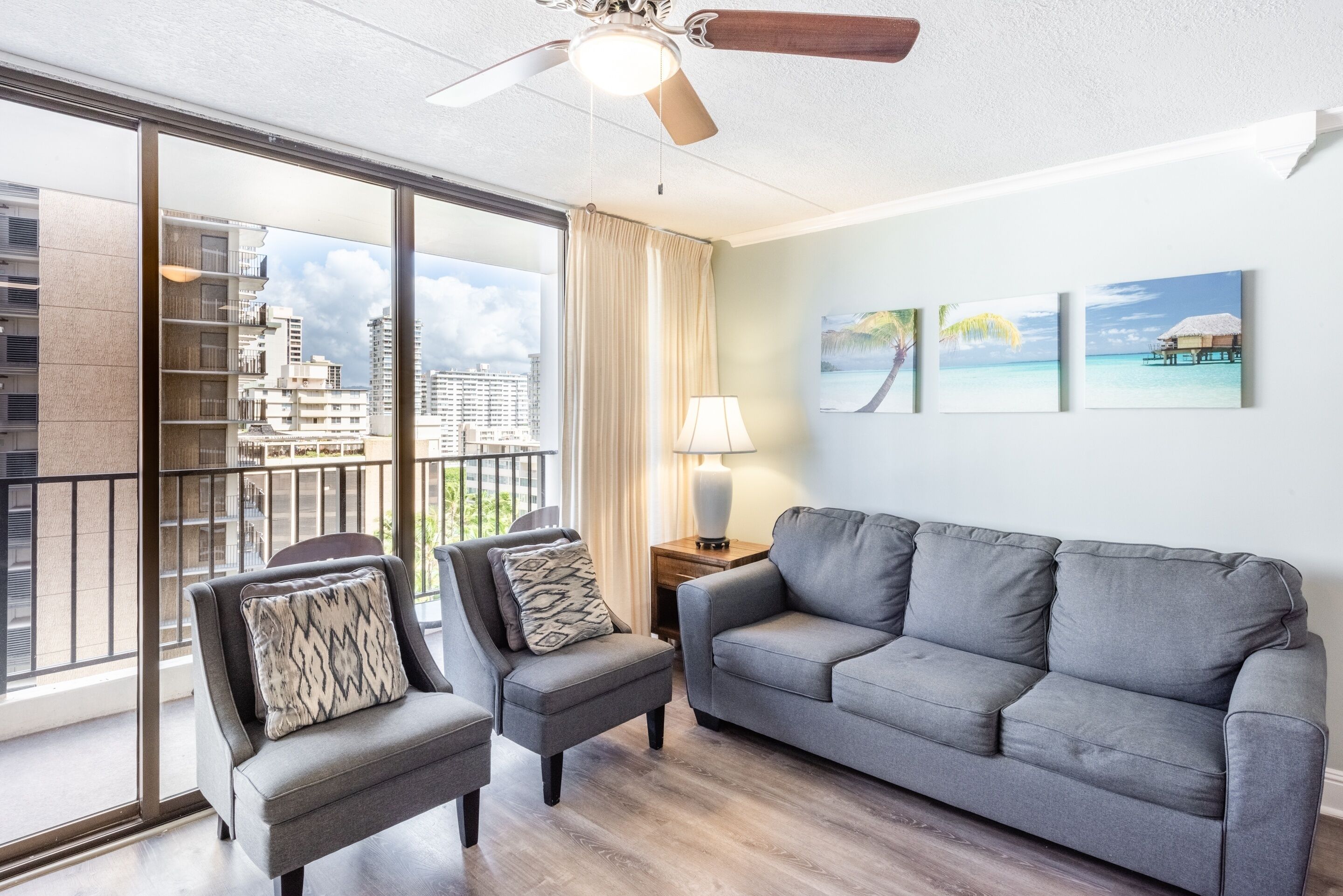 Ein modernes Wohnzimmer mit einem grauen Sofa, zwei Sesseln, einem Deckenventilator und Blick auf eine Stadtkulisse durch große Fenster.