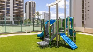Un terrain de jeux avec des toboggans et des structures d’escalade, entouré d’un court de tennis et d’immeubles de grande hauteur.