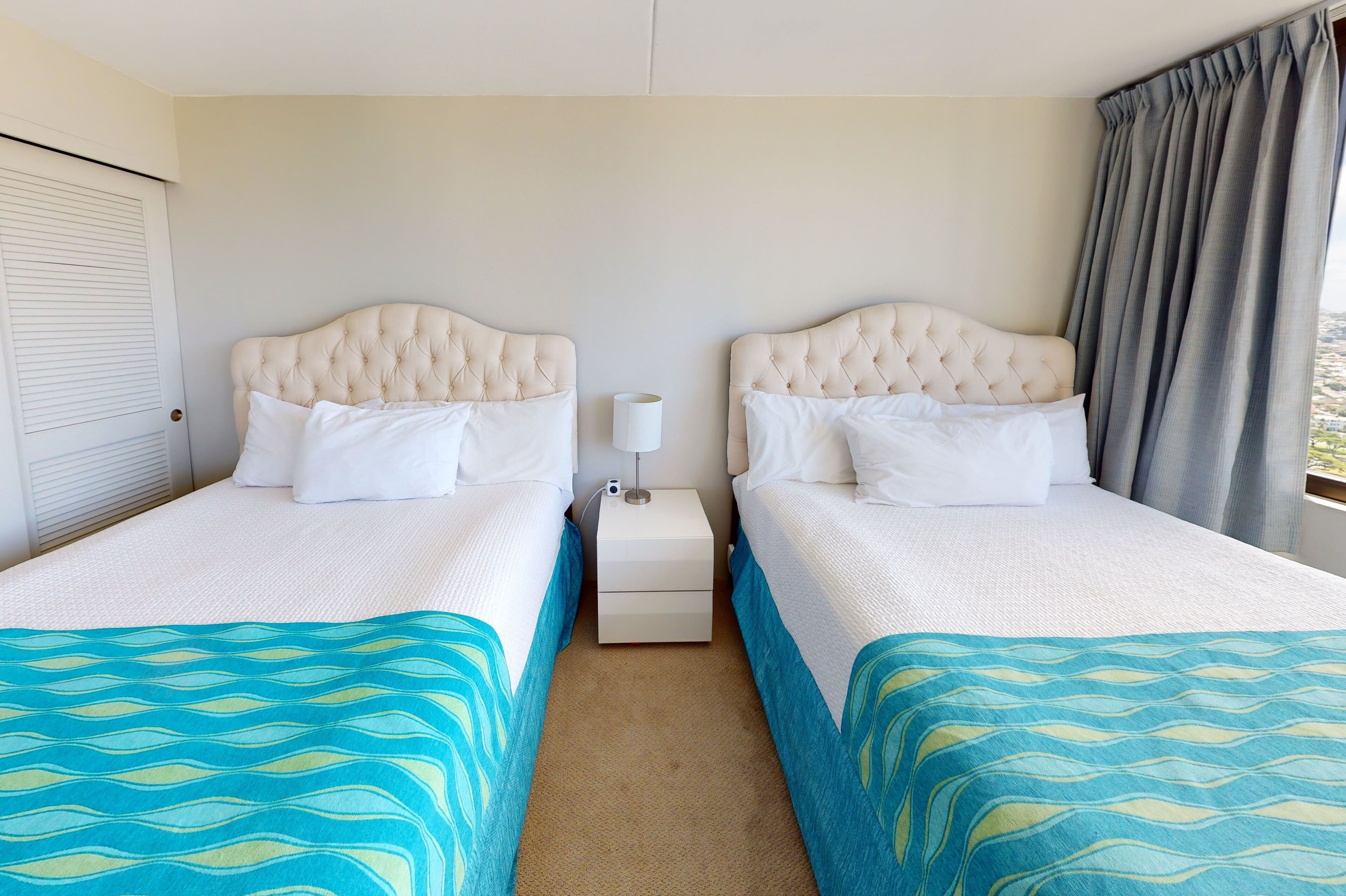 Une chambre d’hôtel avec deux lits, chacun doté d  ’une tête de lit capitonnée, de draps blancs et d’une couvre-lit à motifs bleus et verts. Une petite table de chevet avec une lampe se trouve entre les deux lits.
