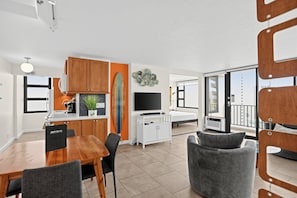 A modern kitchen with wooden cabinets, a dining table with chairs, a TV, and a balcony with a view. - Artsy 32nd Floor Condo with Modern Furnishings & Gorgeous Ocean Views (Honolulu)