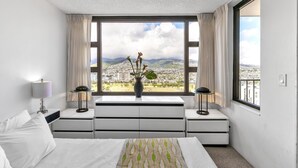 Una habitación de hotel con una cama, una mesita de noche con lámpara, un aparador y una ventana que ofrece vistas a la ciudad y a las montañas.
