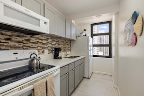 A modern kitchen with a built-in microwave, stove, and coffee maker, a window with a view of buildings, and a decorative fish wall art piece. - Spacious 14th Floor Corner Suite, Partial Diamond Head and Ocean Views, FREE Parking! (Honolulu)
