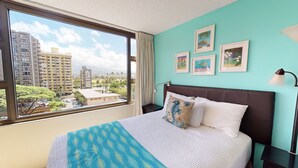 A hotel room with a large window, a bed with a patterned blanket, a headboard, and a nightstand with a lamp.