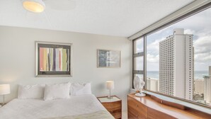 A hotel room with a large bed, a dresser, a nightstand, and a view of tall buildings. - High Rise Ocean View Waikiki Condo, FREE Parking & Wi-Fi (Honolulu)
