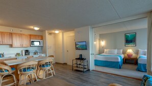 A compact hotel room with a kitchenette, a dining area with wicker chairs, a bed with blue bedding, and a painting of a beach scene. - Updated Waikiki Condo with Mountain Views - 22nd floor, Free parking & WiFi (Honolulu)