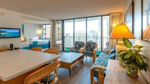 A hotel room with a balcony, a dining area with wicker furniture, a sofa with a blue and green patterned cover, and a large window with a view of buildings. - Updated Waikiki Condo with Mountain Views - 22nd floor, Free parking & WiFi (Honolulu)