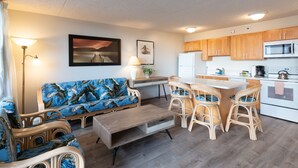 A modern living room with a sofa, coffee table, and wicker chairs. A kitchen with wooden cabinets and white appliances is visible. - Updated Waikiki Condo with Mountain Views - 22nd floor, Free parking & WiFi (Honolulu)