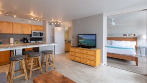 A modern kitchen with a central island, bar stools, a television, and a bed with a view. - High Level Waikiki Condo - Enjoy Ocean Views From Your Private Lanai! (Honolulu)