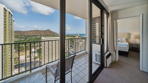 Un balcón con vista a un paisaje urbano y montañas, una cama con una mesita de noche, y una ventana con cortinas.