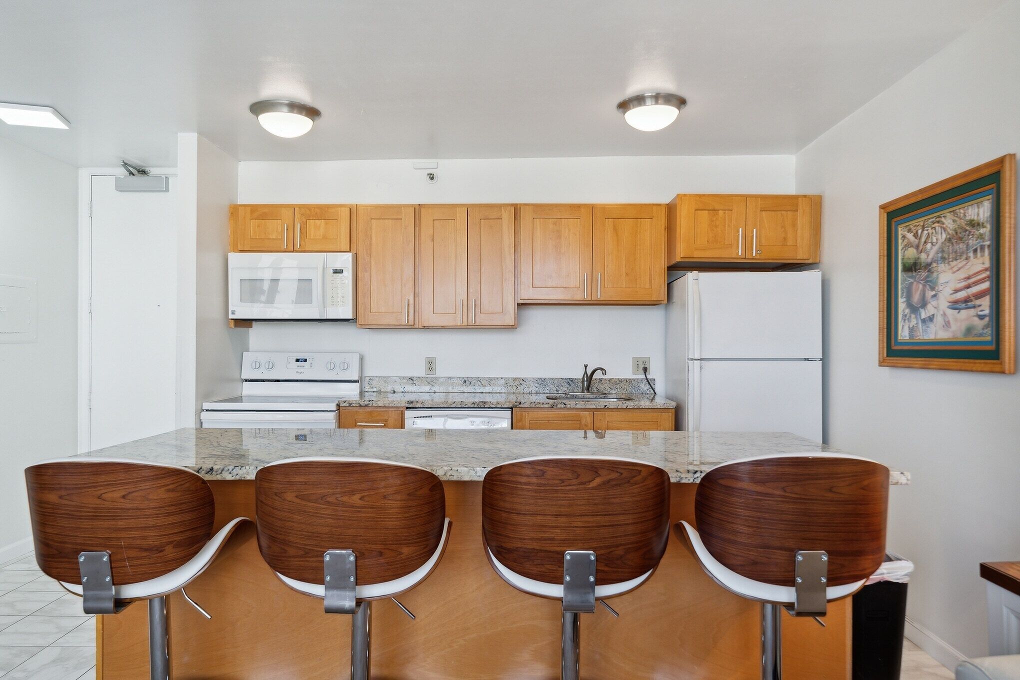 Eine moderne Küche mit Holzschränken, einem weißen Kühlschrank, einer Mikrowelle und einer Arbeitsplatte aus Granit. Drei Barhocker mit Metallbeinen sind vorhanden.