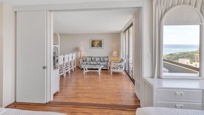 Hotellihuone, jossa on puulattia, valkoiset huonekalut, kukkakuviollinen sohva, ruokailutila tuoleineen, suuri ikkuna, josta on näkymä, ja valkoinen lipasto.