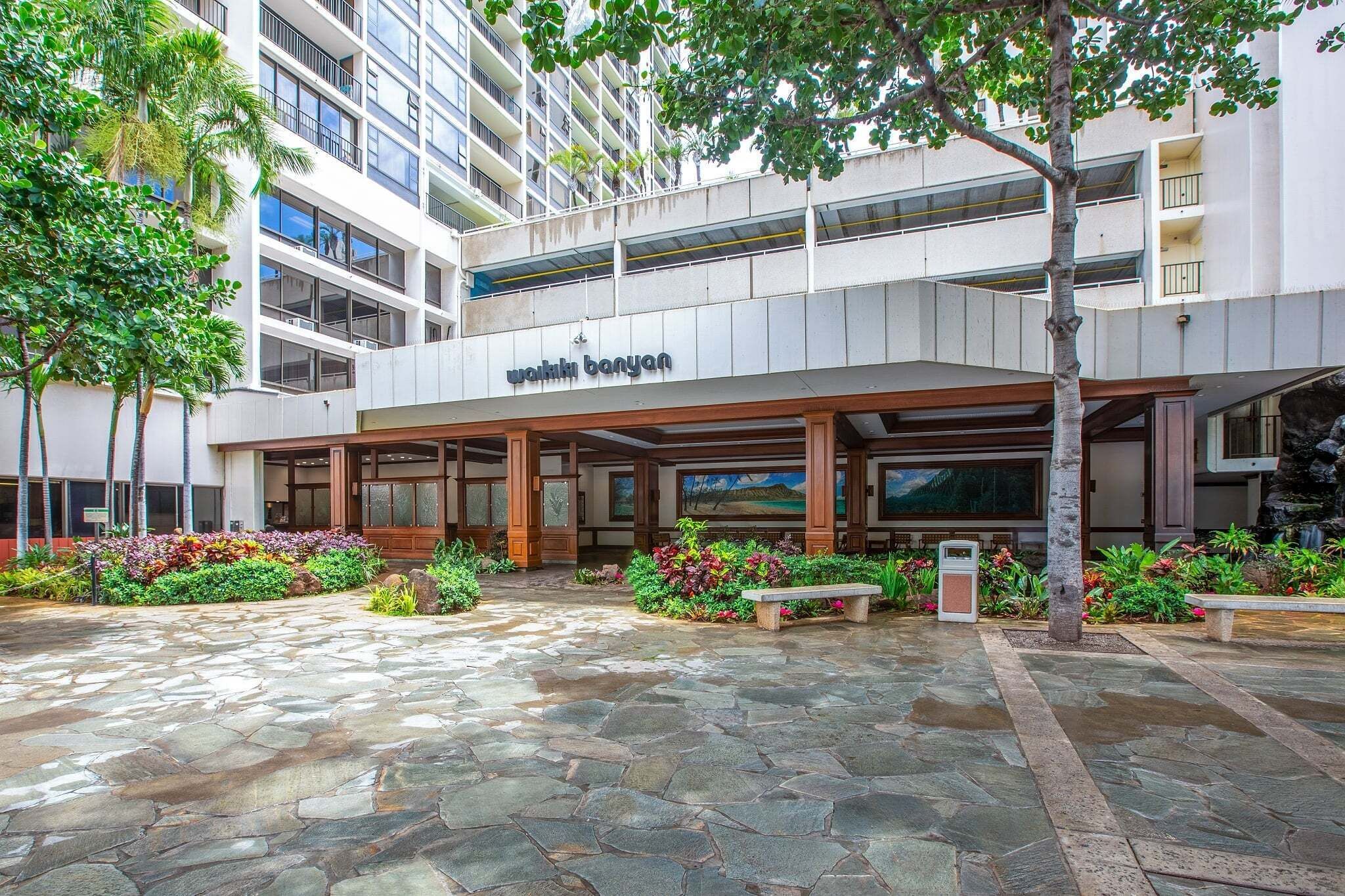 A modern building with a covered entrance, stone-paved courtyard, and lush greenery.