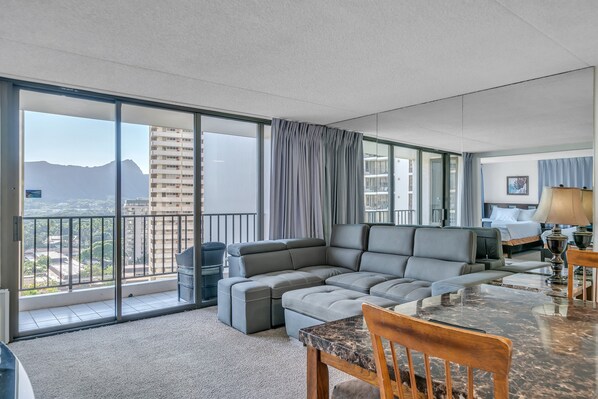 A modern hotel room with a large gray sectional sofa, a dining table with chairs, and a balcony with a view of the city and mountains. - Excellent Diamond Head View Condo - Remodeled, Free Parking! (Honolulu)