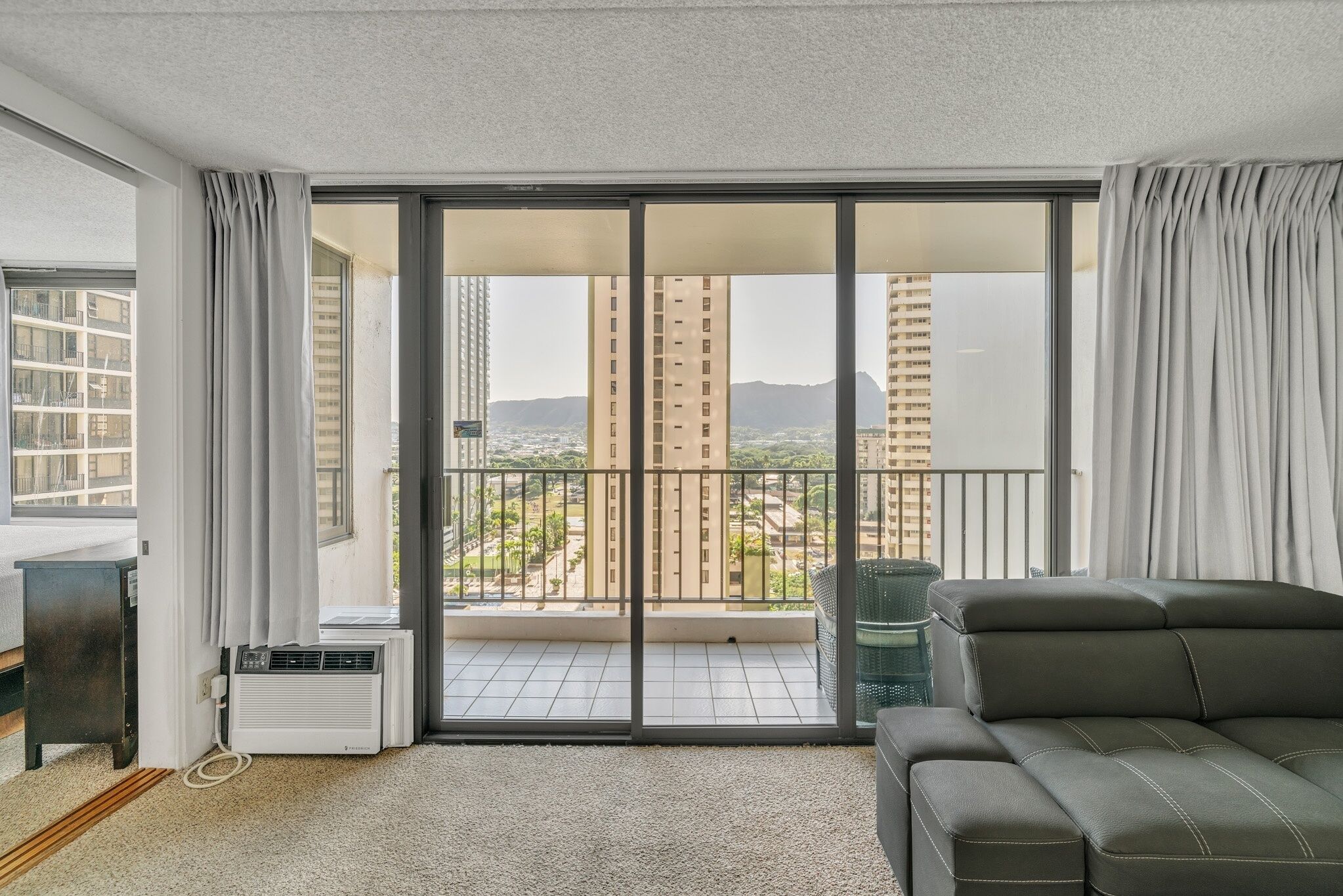 A modern living room with a grey sofa, a small table, and a sliding glass door leading to a balcony with a city view.