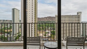 A balcony with two chairs and a small table, overlooking a cityscape with tall buildings and a distant mountain range. - Gorgeous High Rise Waikiki Condo with Ocean and Diamond Head Views (Honolulu)