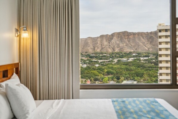 A hotel room with a bed, curtains, and a view of a mountainous landscape and buildings.