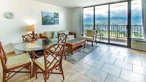 A spacious living area with a glass table, wooden chairs, and a sofa. A large window offers a view of mountains. - Deluxe Panoramic Mountain View Condo - 37th Floor, Free parking & Wifi (Honolulu)