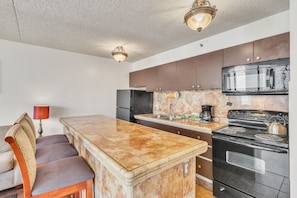 A kitchen with a marble countertop, stainless steel appliances, and a dining area with a chair and lamp. - 19th Floor Condo with Lanai & Sea Views - FREE Parking! (Honolulu)