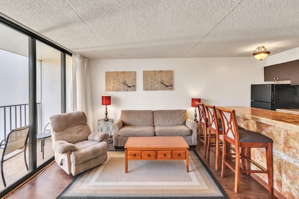 A living room with a sofa, armchair, coffee table, and a dining area with chairs. There are two wall-mounted artworks, a kitchen area with a refrigerator, and a balcony with a chair and table. - 19th Floor Condo with Lanai & Sea Views - FREE Parking! (Honolulu)